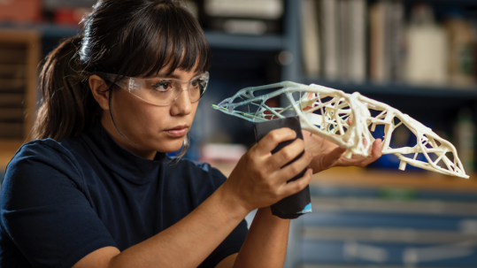 Woman wearing protective goggles looking at a miniature structure in a her hand