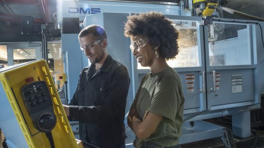 Man in overalls in a factory with a woman overseeing both wearing protective goggles