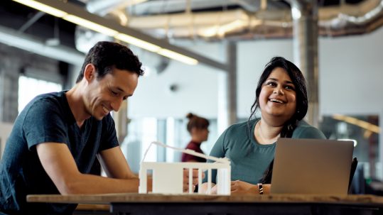 Two people working with AutoCAD on an open laptop at a desk while laughing