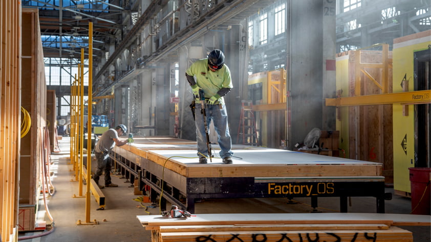 Two workers build a modular unit in a well-lit warehouse. One uses a power tool; the unit is labeled “Factory OS.”