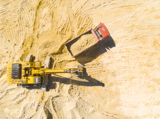 Aerial view of a yellow excavator loading dirt into a red dump truck at a sandy construction site