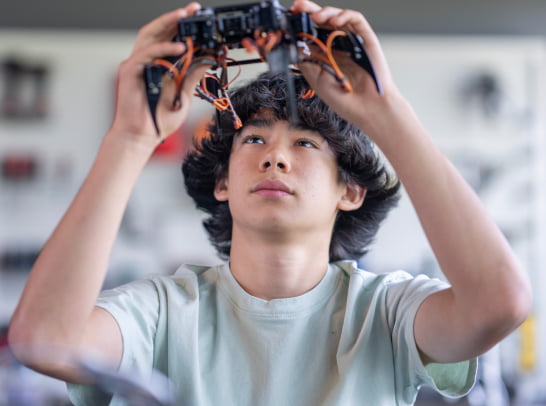 Teenager studies a mechanical insect-like robot in a robotics lab, holding it up to observe its wiring and build