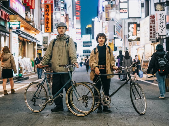 Amid bright signage and dense foot traffic, two cyclists pose in what appears to be a popular nightlife district in Japan