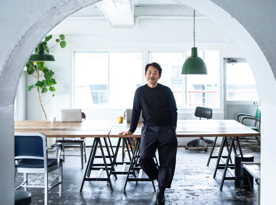 A man stands in a bright industrial loft framed by a white concrete arch, with raw floors, mismatched chairs, and green pendant lights accenting a long wood table