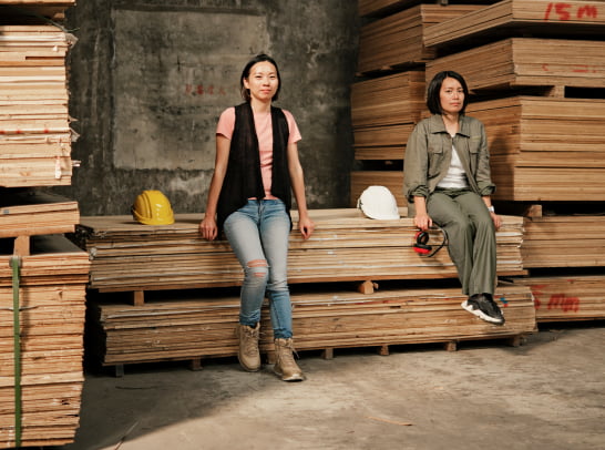 Two women sit confidently on a stack of plywood sheets inside an industrial warehouse, surrounded by neatly piled timber and rough concrete walls, with yellow and white hard hats beside them