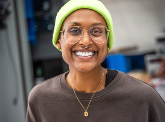 In a candid moment, a person with a wide smile, vibrant green beanie, and small gold necklace stands relaxed in a modern, organized workspace with shelves and tools in the background