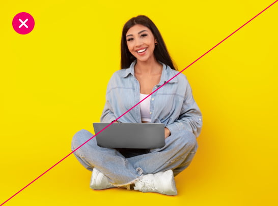 A woman sits cross-legged against a bright yellow background, her laptop in her lap. The image is marked with a red X.
