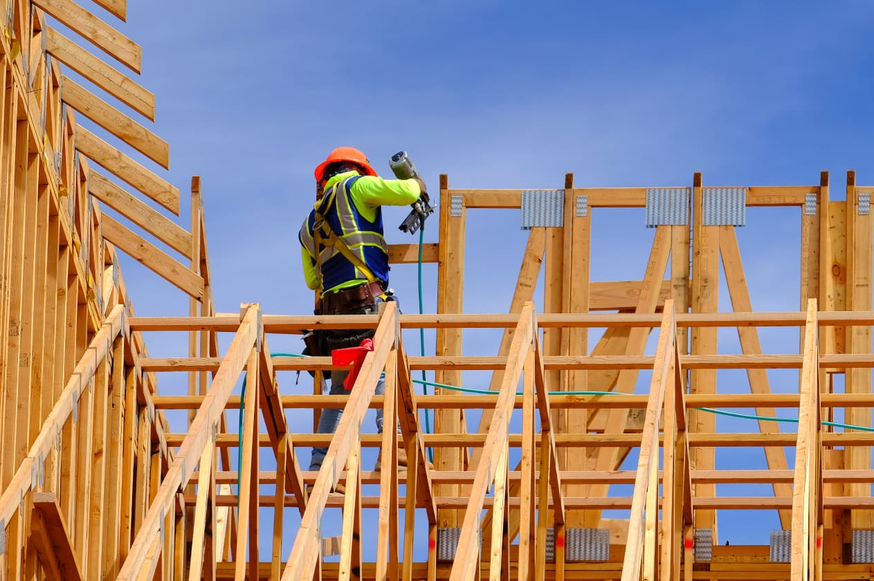 A construction worker in a safety harness uses a nail gun while standing on a partially built wooden roof frame