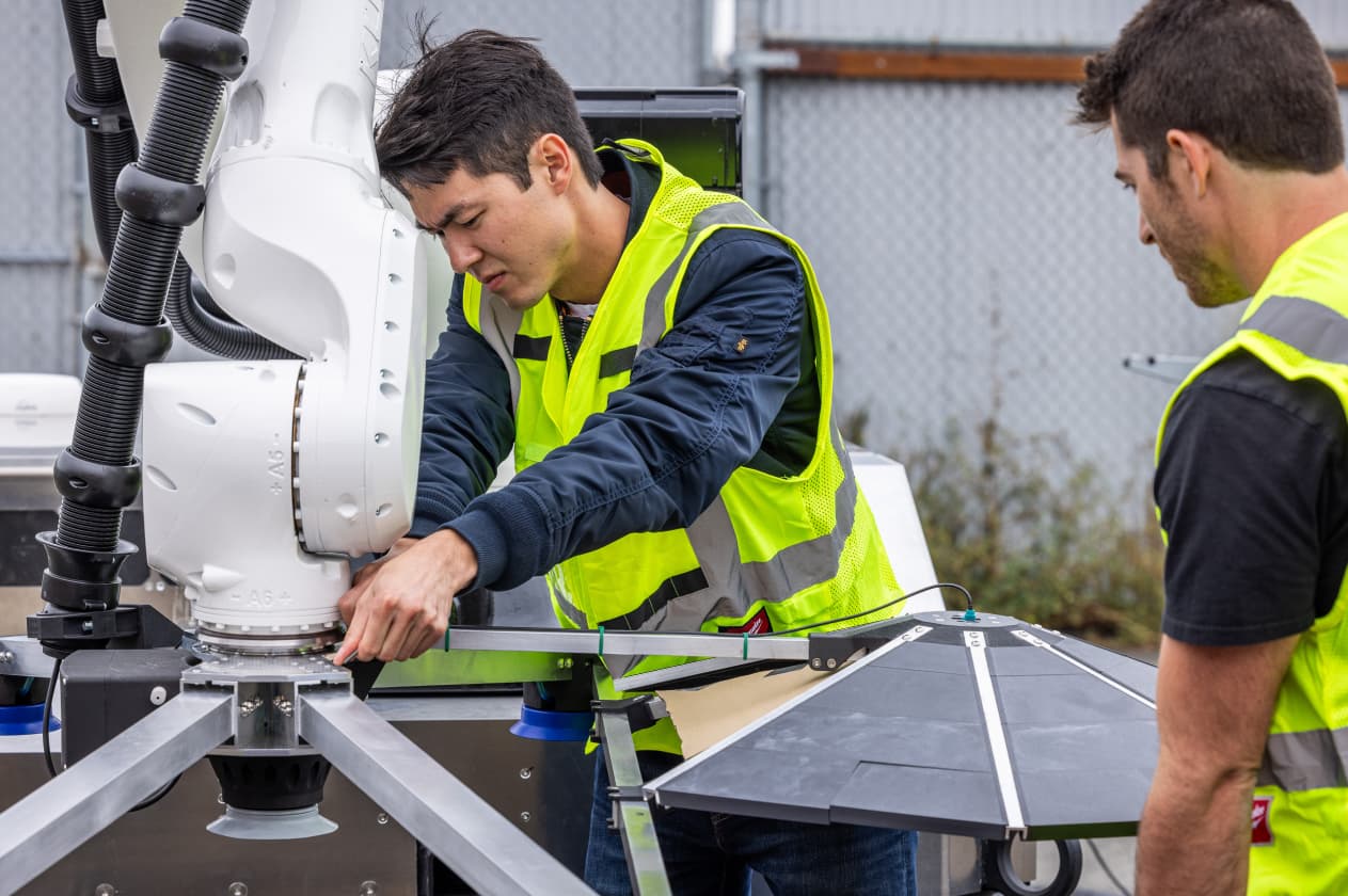 A technician adjusts a robotic arm outdoors while another worker observes, both wearing safety vests