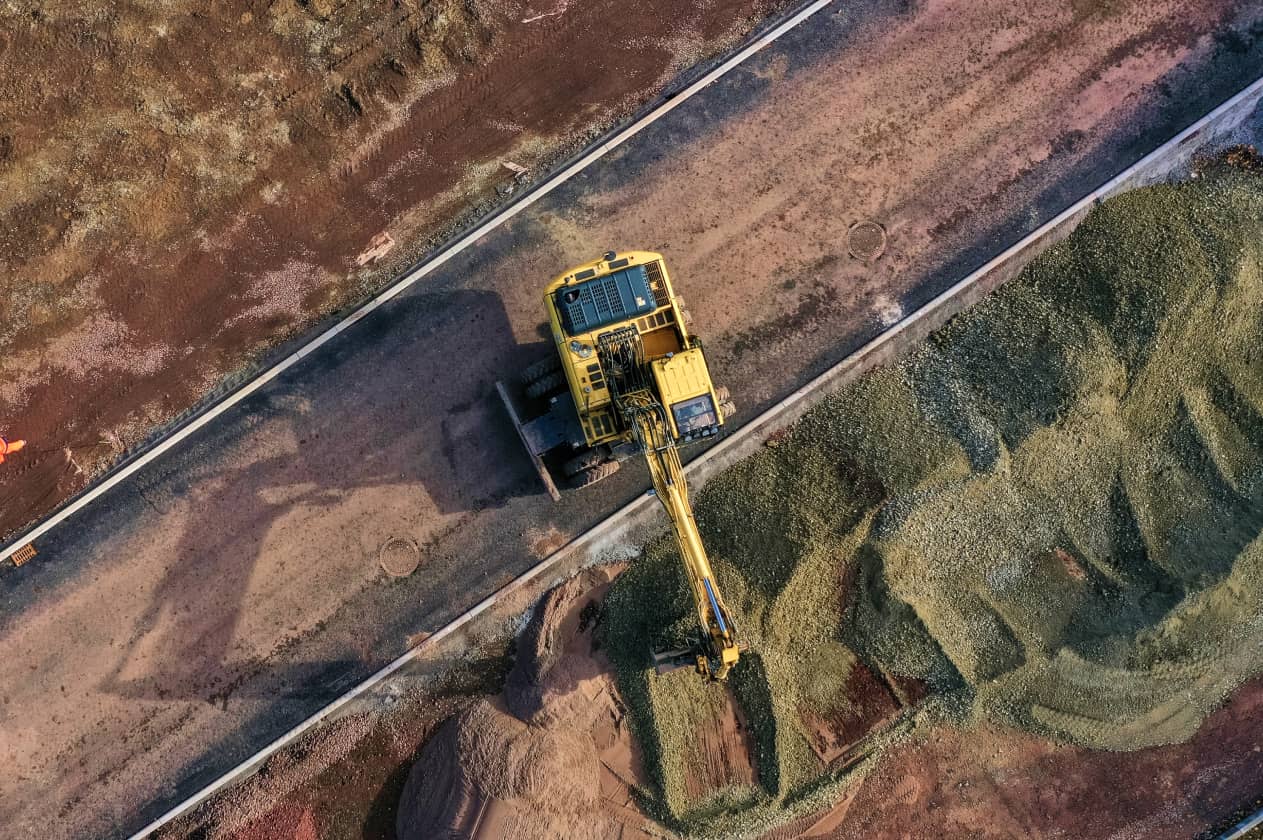 Overhead view of a large yellow excavator moving piles of gravel along a dirt construction site, casting a sharp shadow on the roadway below