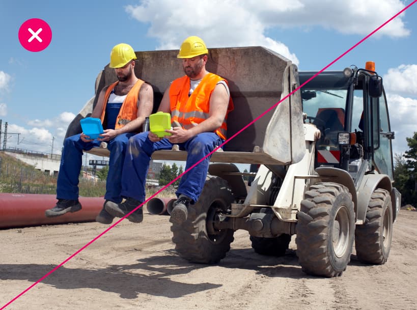 Construction scene showing two men in reflective vests and hard hats having lunch inside a loader bucket, highlighting improper safety practices. Marked with a red X.