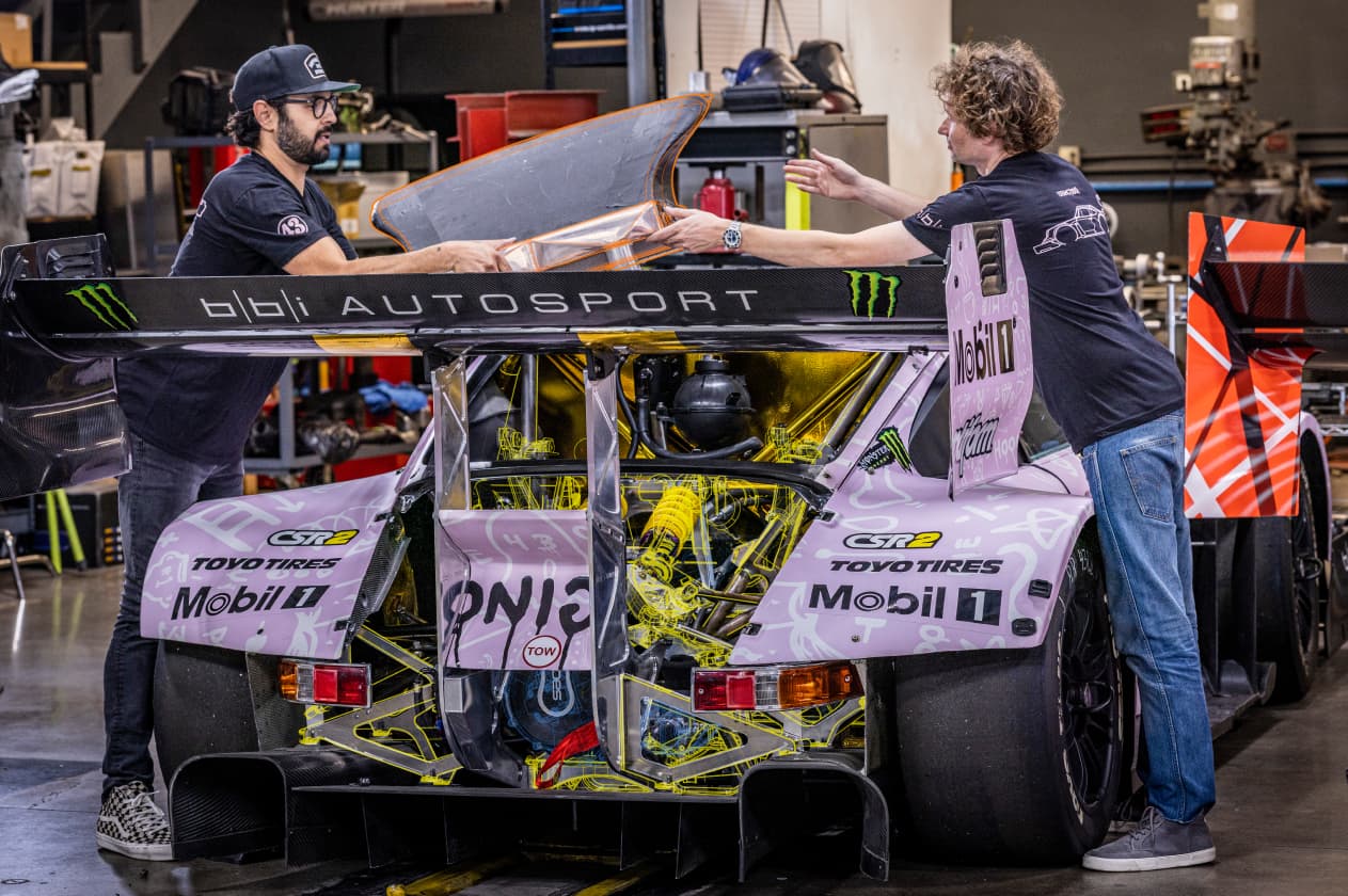 In a garage workshop, two mechanics work on a race car, its interior frame digitally highlighted in bright yellow