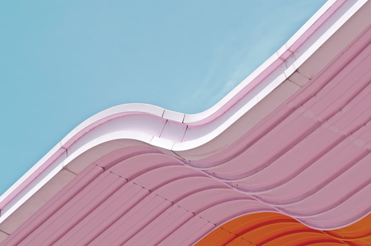 Abstract close-up photo of a curving, undulating roof, painted in pastel pink and bright orange, set against a blue sky
