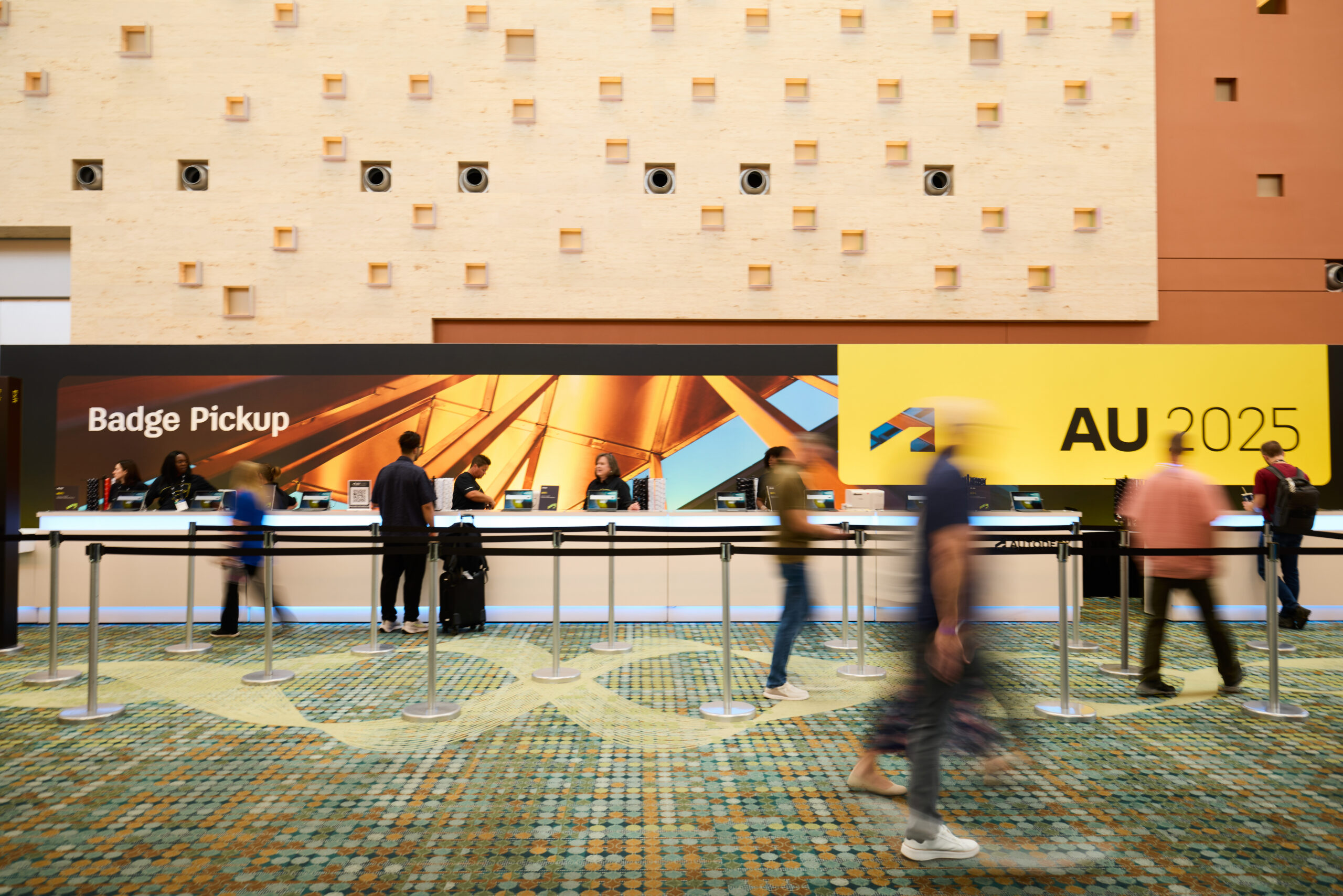 A photograph of the registration counter of Autodesk University 2025 with a large abstract photo in the background and the words "Badge Pickup" and "AU2025" overlaying the graphic